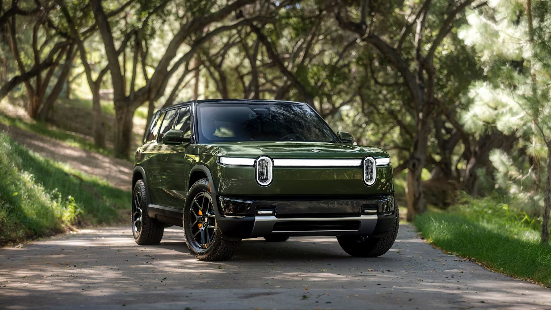 Green Rivian R2 electric SUV, front three-quarter view on a tree-lined road
