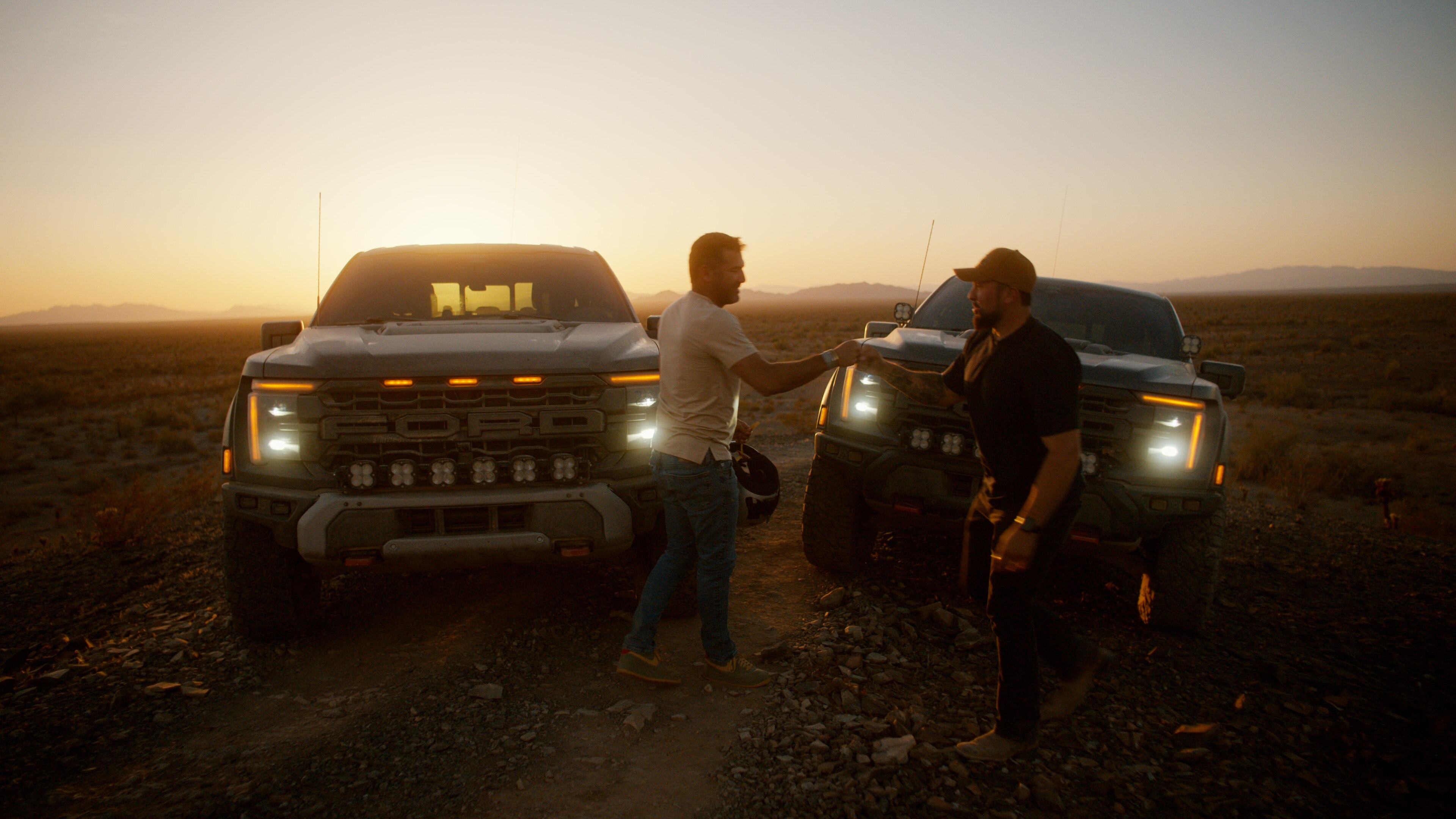 Two Ford F-150 Raptor trucks at sunset with two drivers in the desert