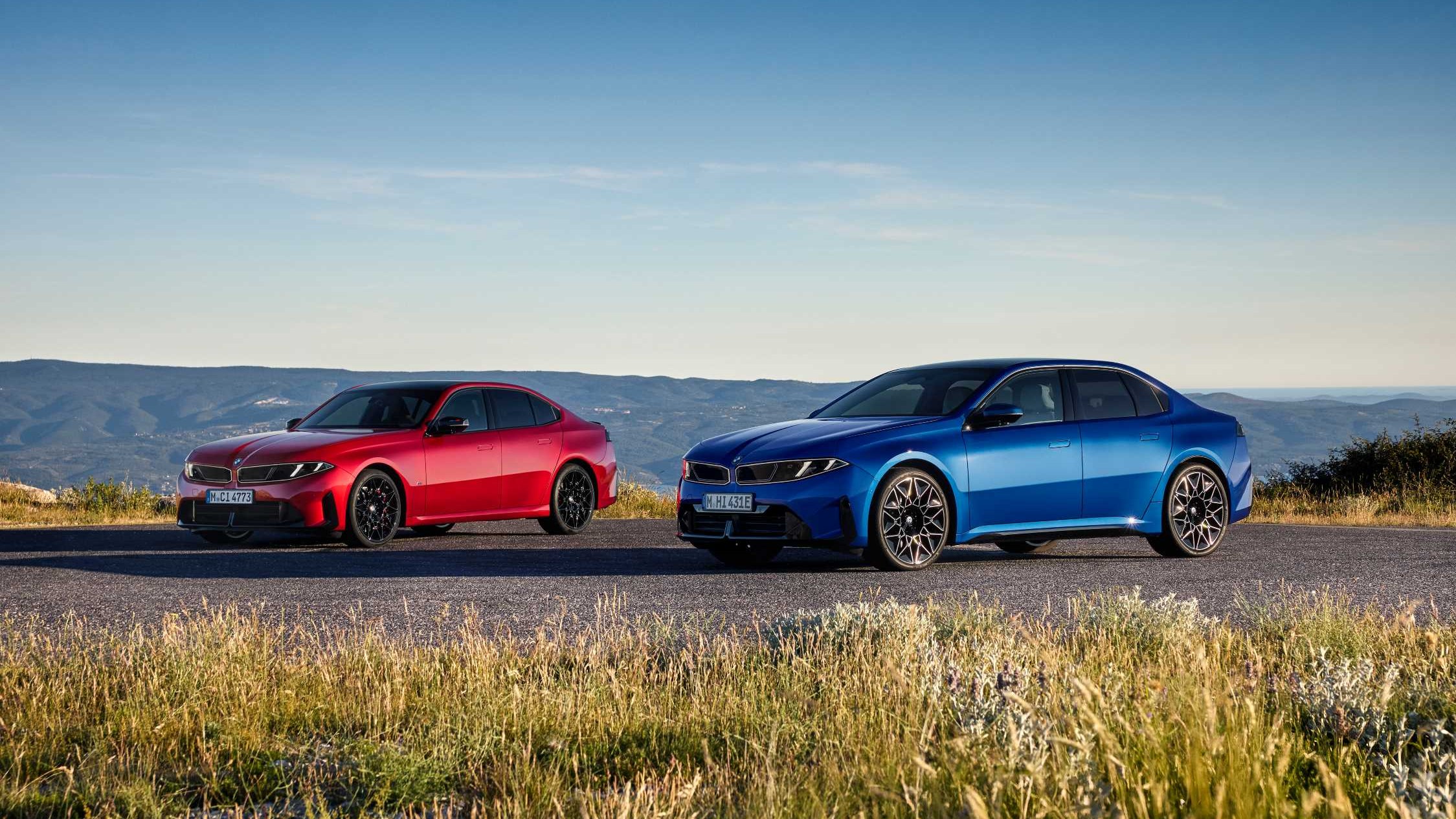 Two new BMW 3 Series sedans, one red and one blue, parked on a mountain road