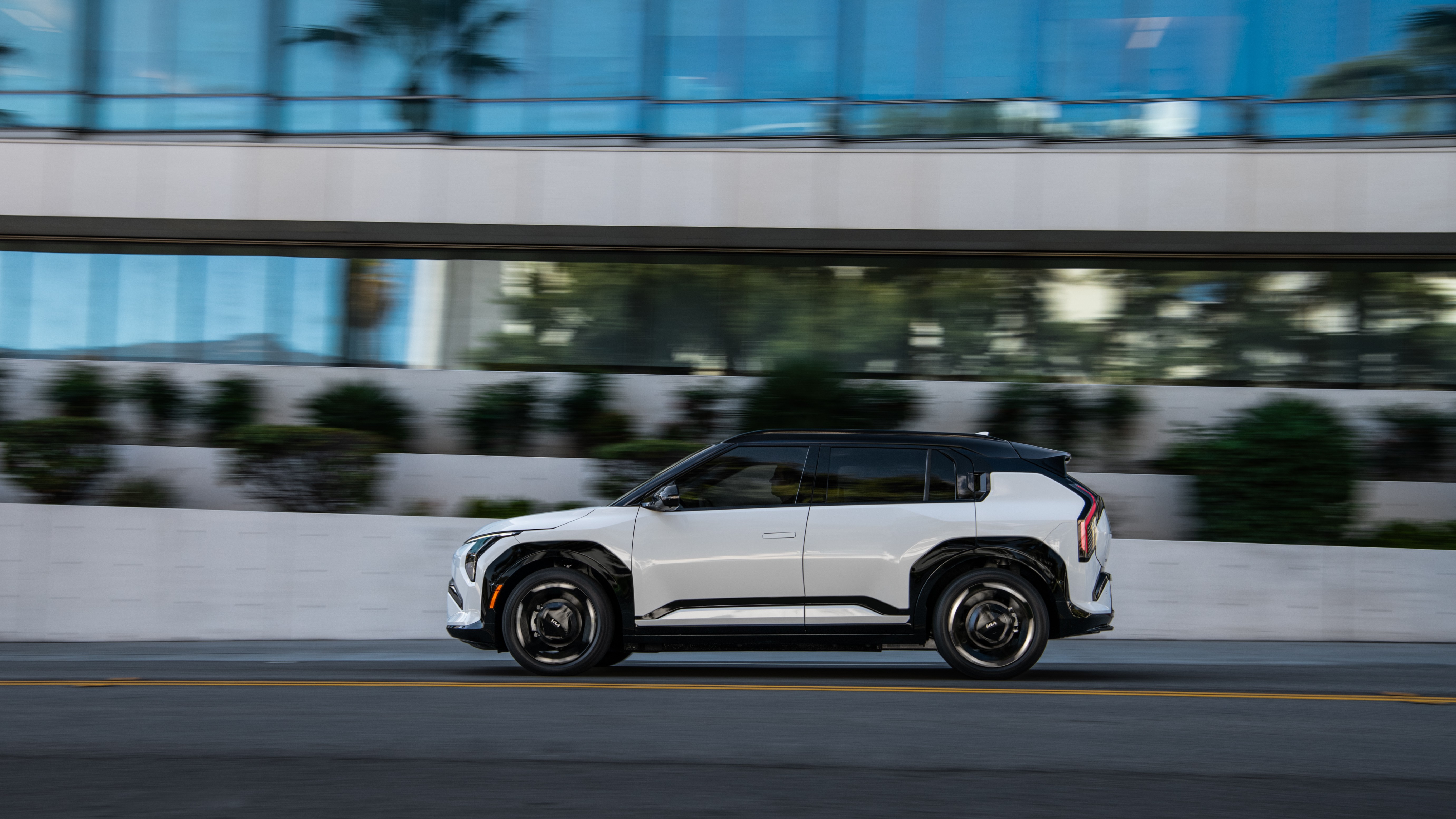 White 2027 Kia EV3 with a black roof driving past a glass office building, side view