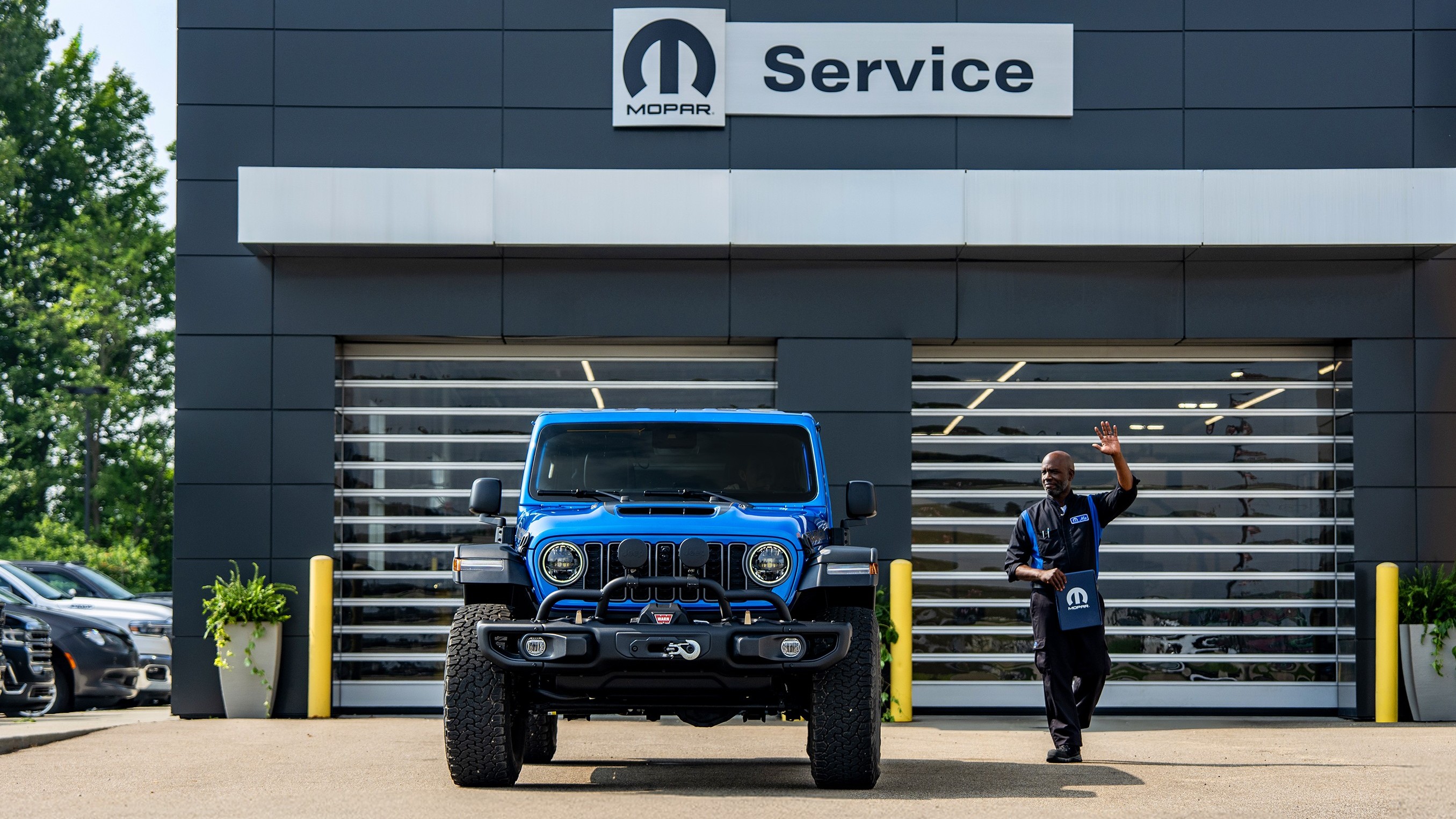 Blue Jeep Wrangler leaving a Mopar Service building as a technician waves
