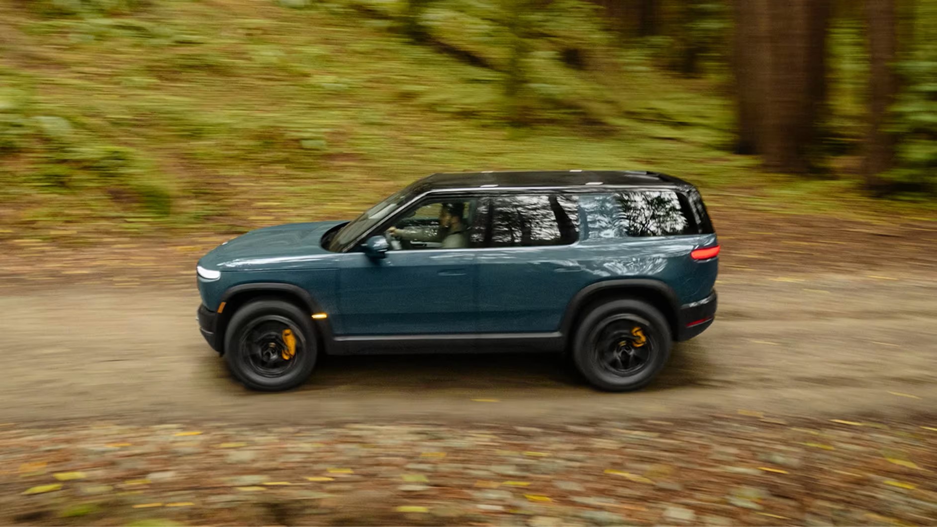 Blue Rivian R2 driving on a forest dirt road