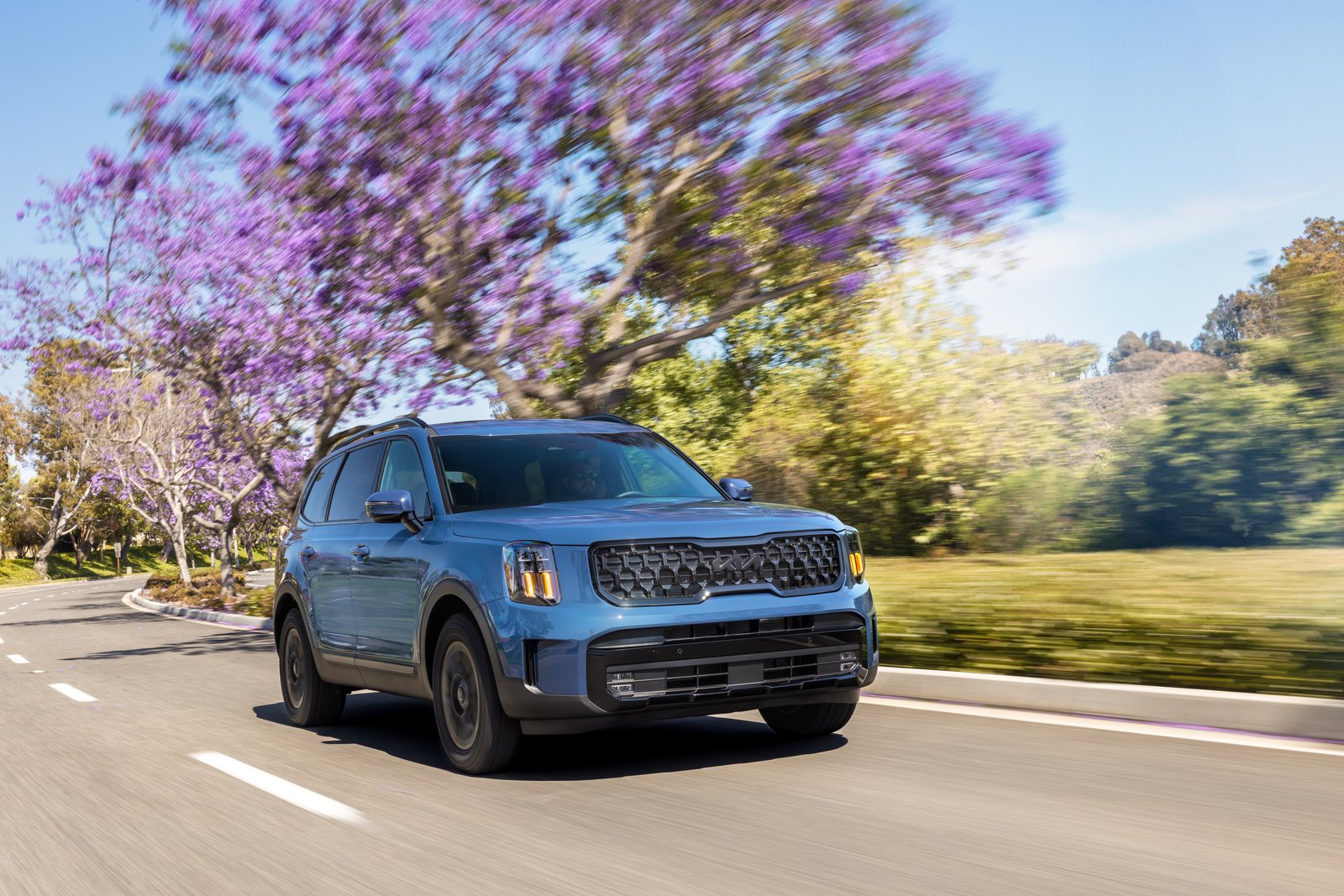 Blue 2025 Kia Telluride driving past blooming jacaranda trees