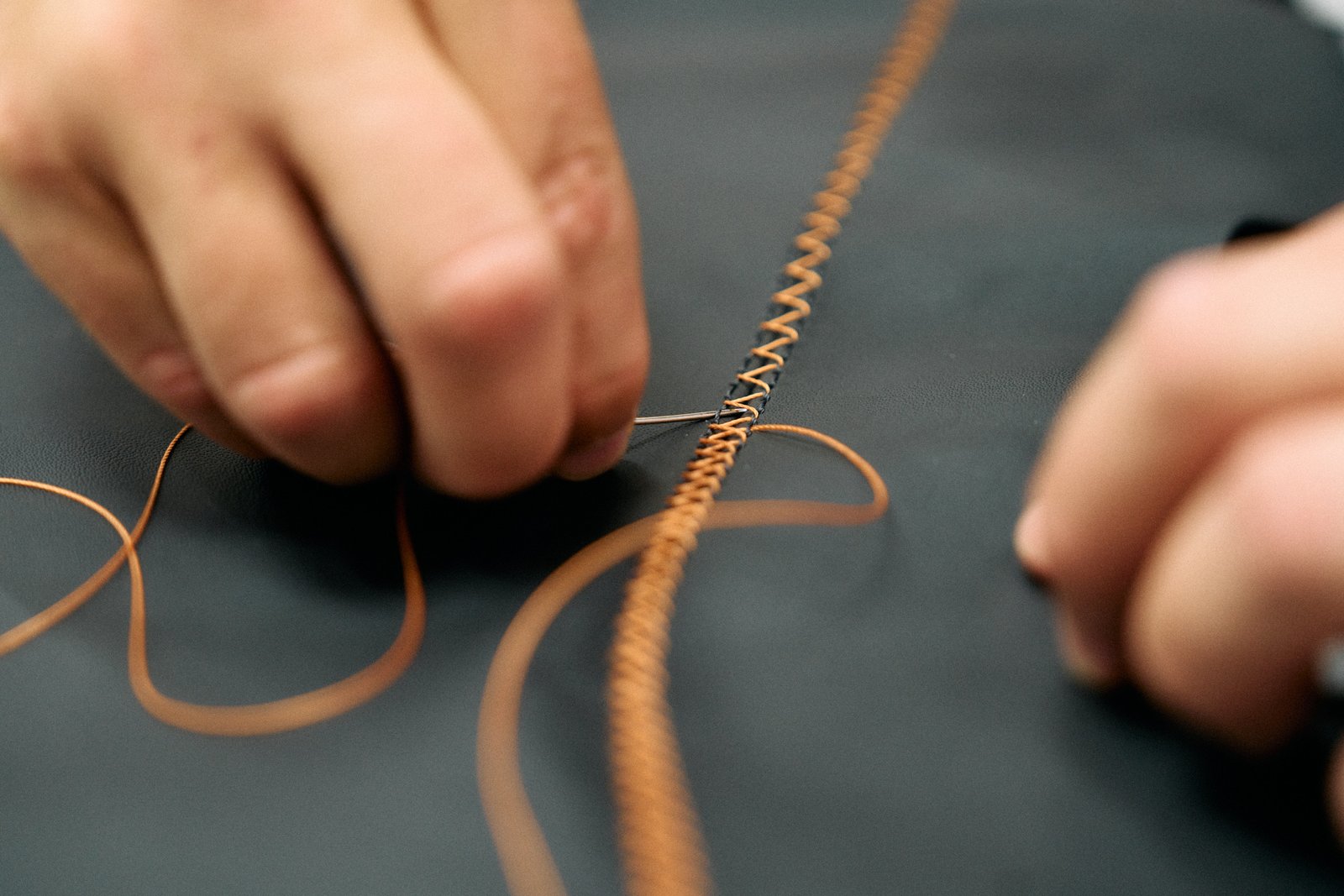 Hands cross-stitching orange thread on dark leather