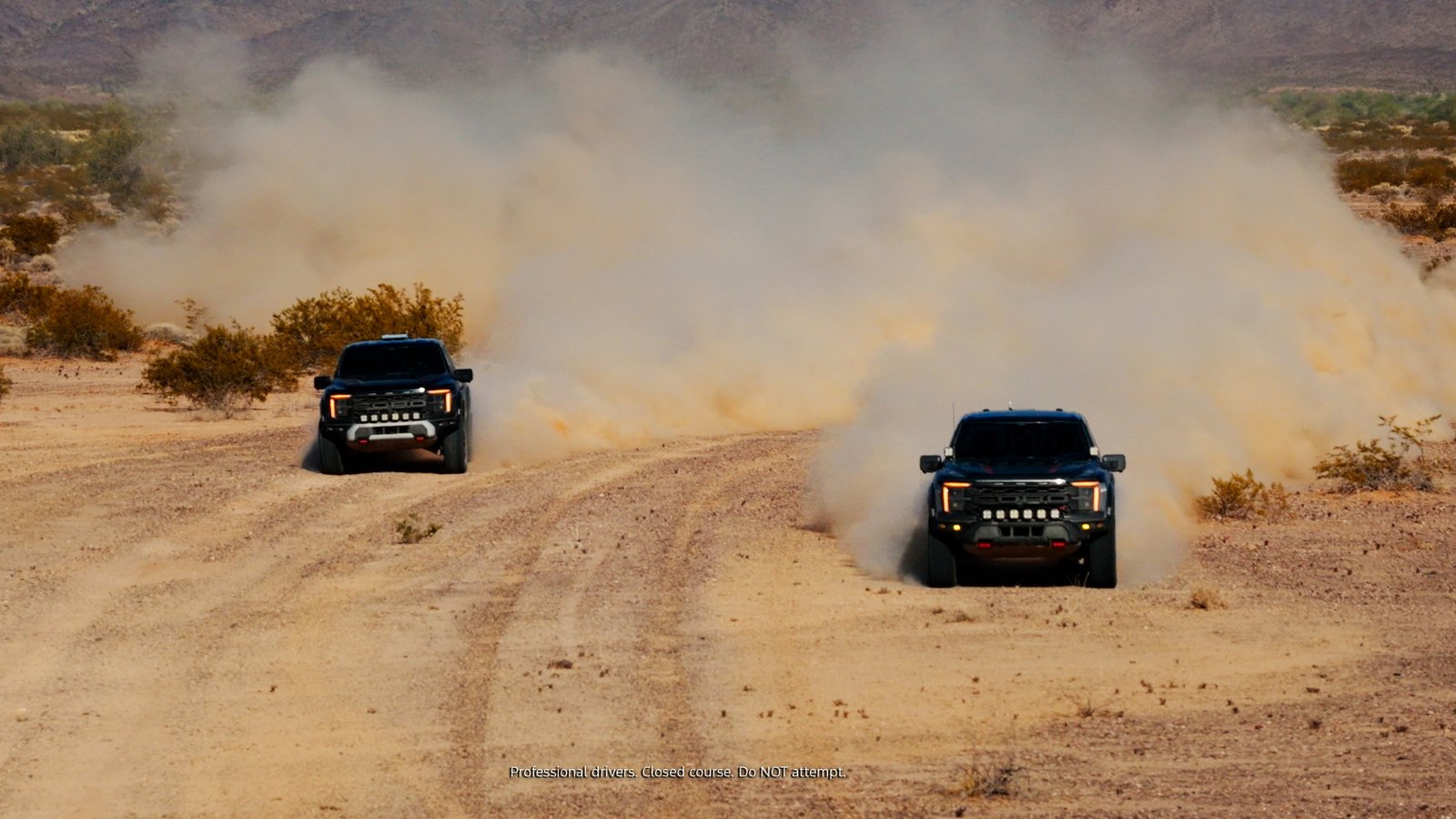 Two black Ford F-150 Raptors driving side by side through desert dust