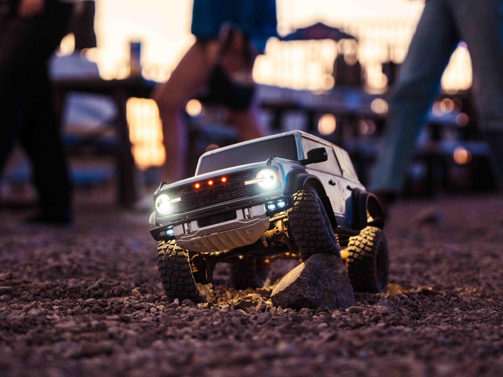 RC-scale Ford Bronco with headlights on crawling over gravel at dusk