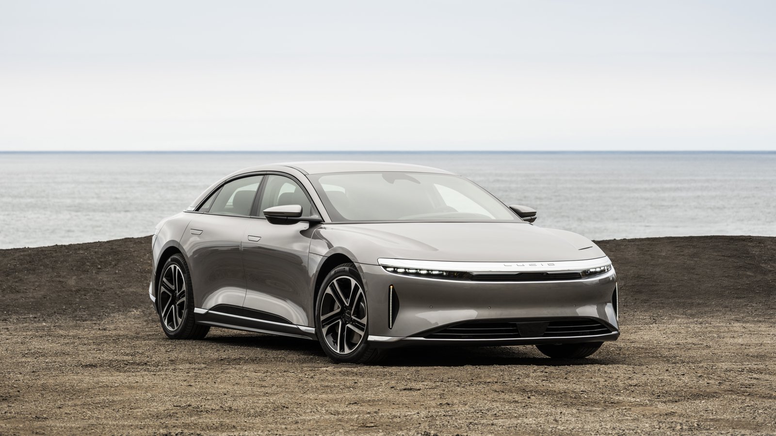 Silver-gray Lucid Air sedan parked on gravel by the ocean, front three-quarter view