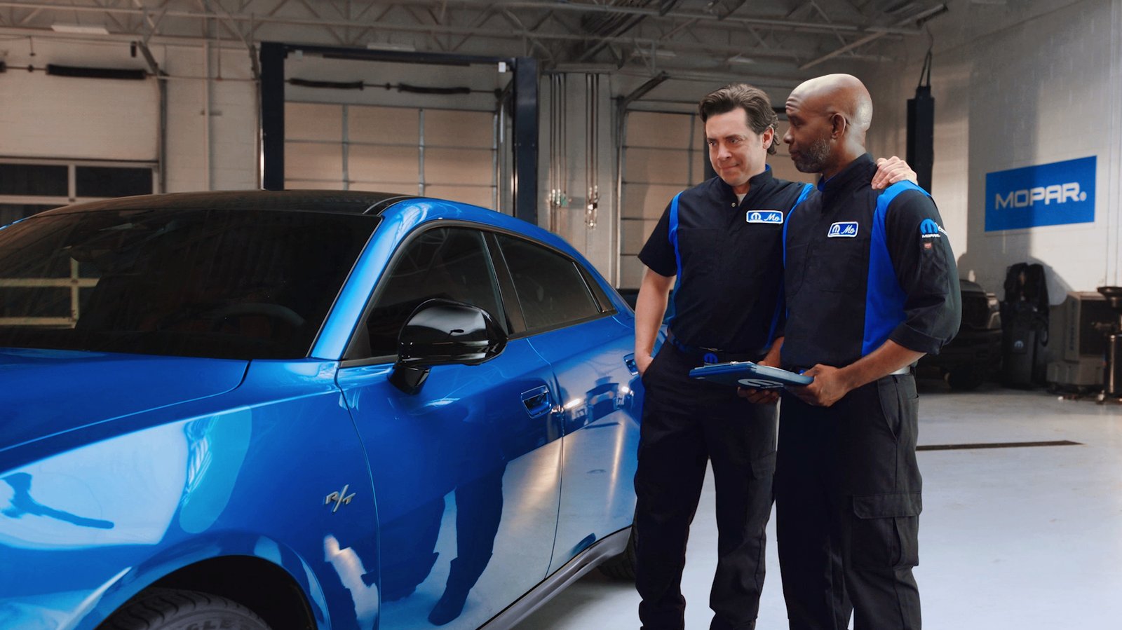 Two Mopar technicians in uniform standing beside a blue Dodge Charger