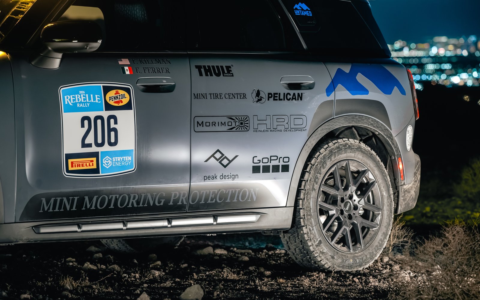 Side of the MINI Countryman rally car showing sponsor decals and all-terrain tires