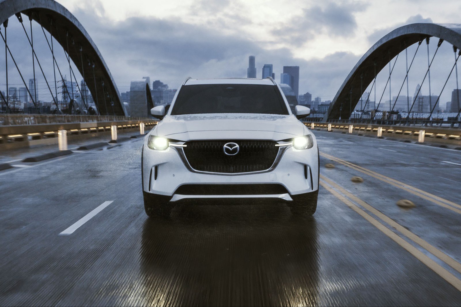 White Mazda CX-90 driving across a wet arched bridge at dusk