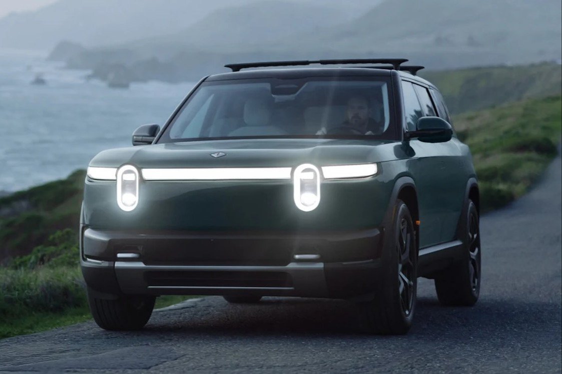 Green Rivian R2 driving on a coastal road with lights on