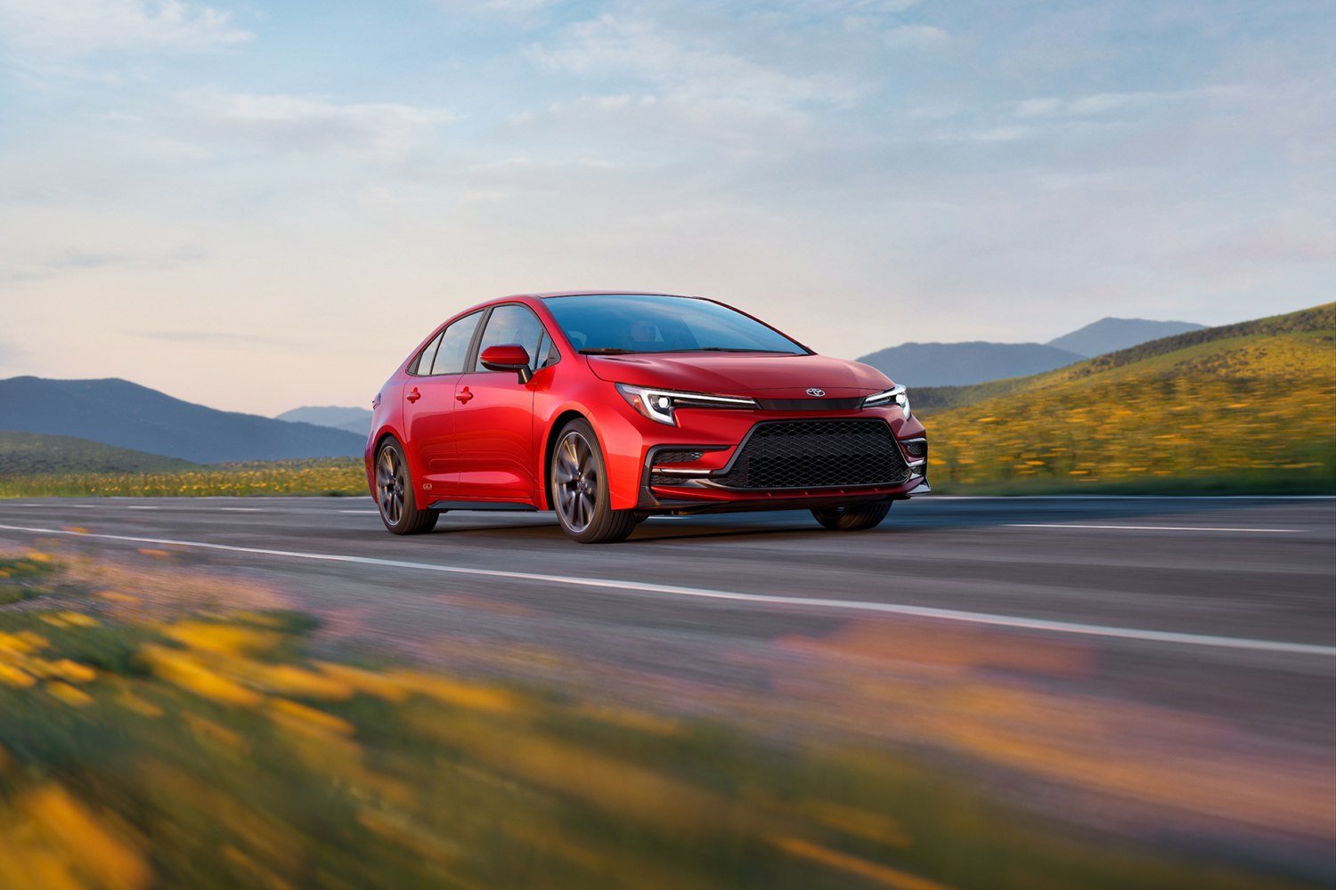 Red 2027 Toyota Corolla sedan driving past hills and wildflowers